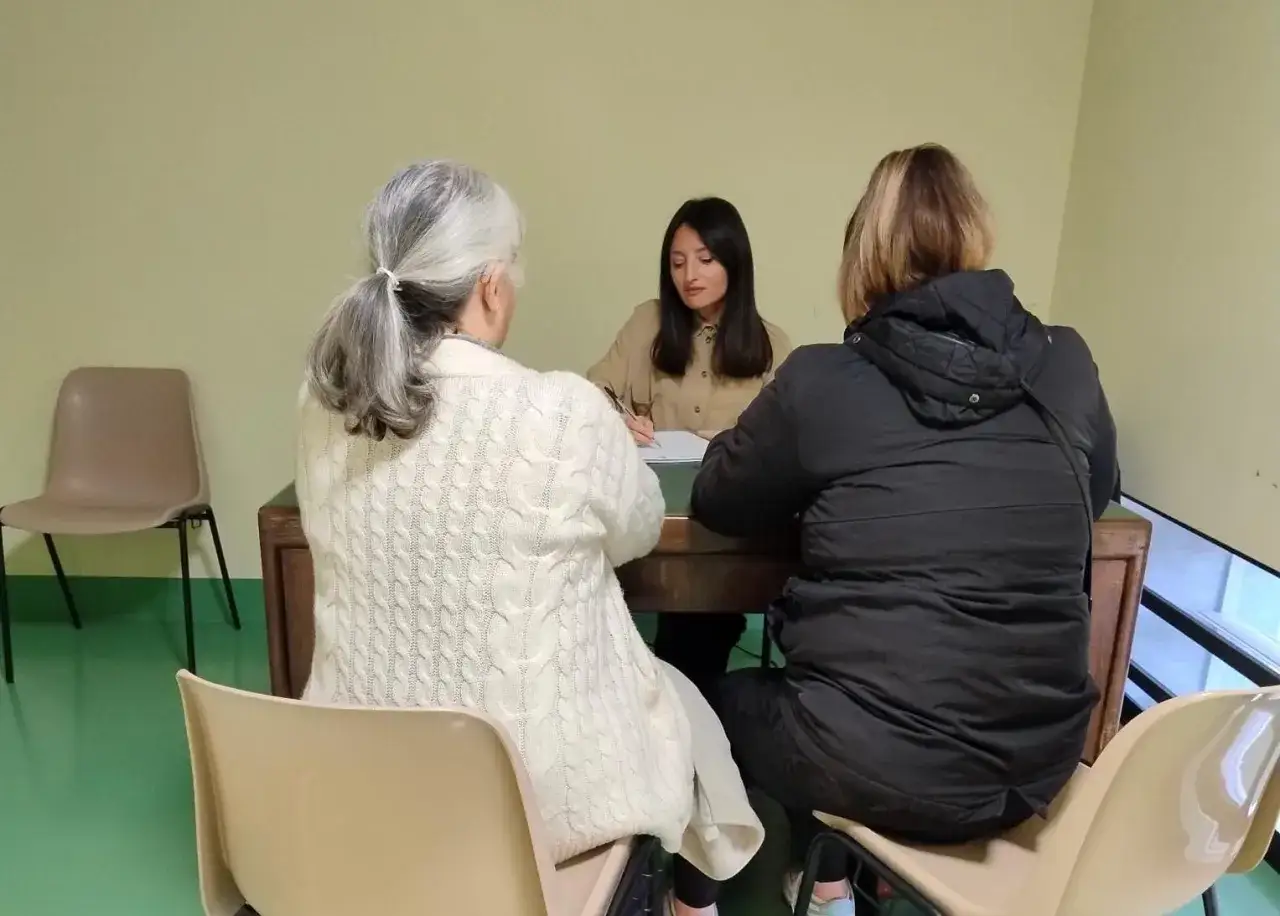 Incontro per pratiche INPS: una donna anziana con un maglione bianco e una donna con una giacca nera parlano con un'impiegata.