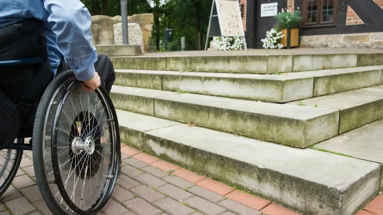 Persona in carrozzina davanti a una scalinata. Le scale rappresentano cosa sono le barriere architettoniche, impedendo l'accesso.